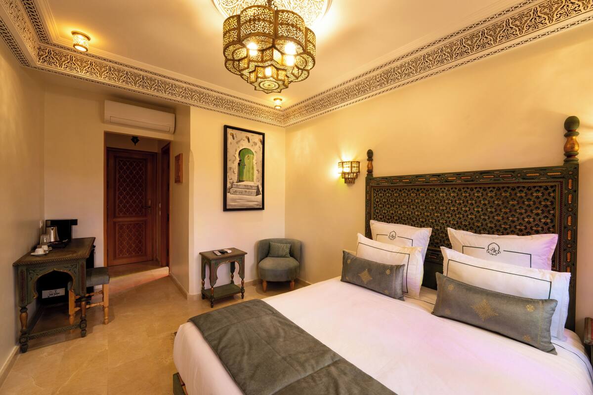 A hotel room with a large bed, a chandelier, a wooden headboard, a small table, a chair, and a framed picture on the wall.