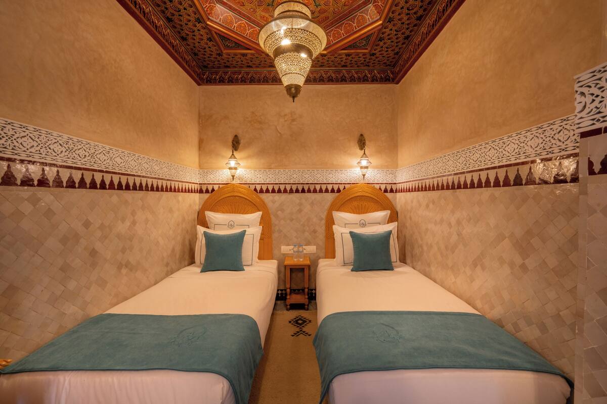 A room with two beds, a chandelier, patterned walls, and a decorative ceiling.