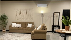 A living room with two sofas, a floor lamp, a wall-mounted air conditioner, and a potted plant. - The serenity studio near route 66 (Webb City)