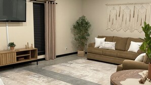 A living room with a sofa, a TV, a plant, and a macrame wall hanging. - The serenity studio near route 66 (Webb City)