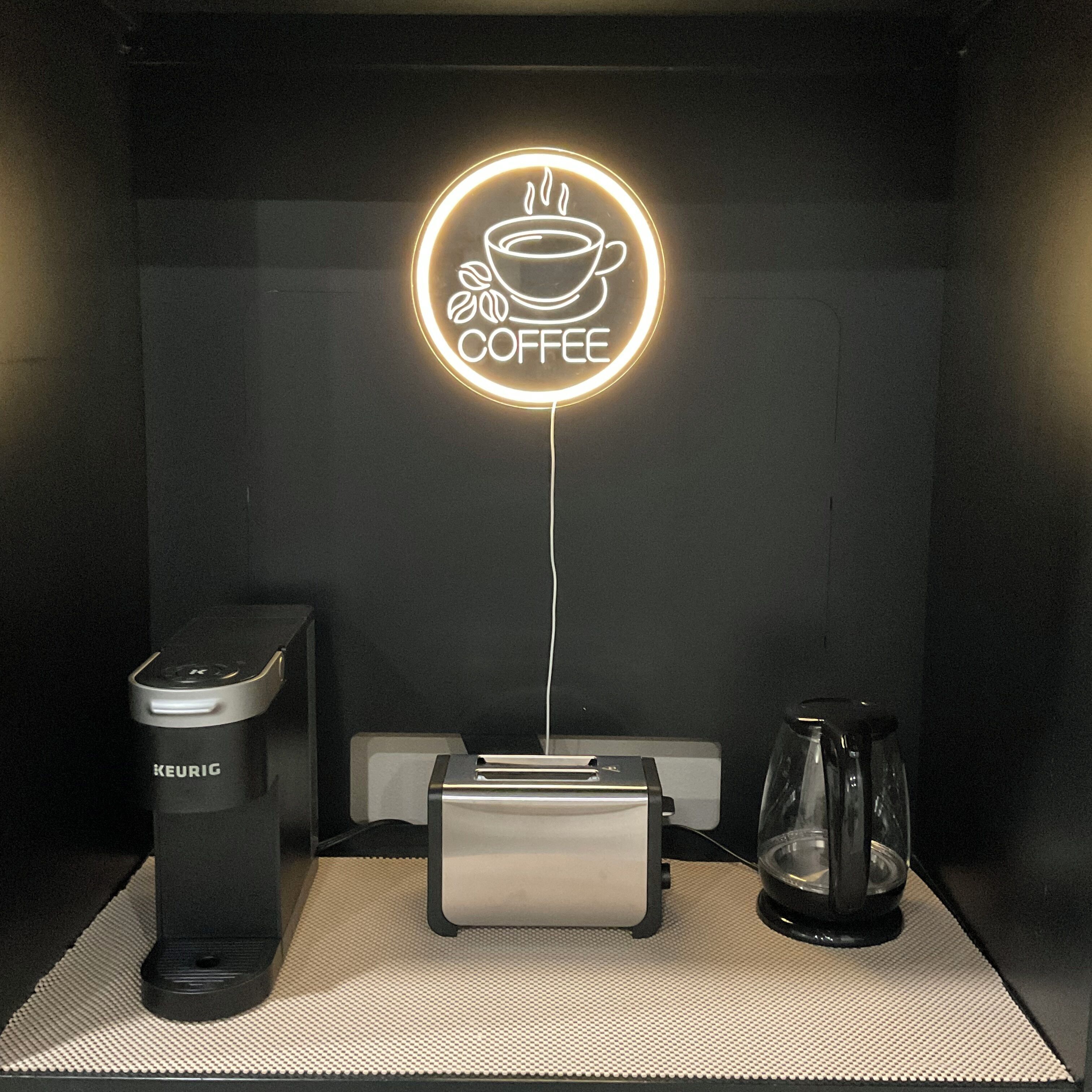 Una macchina da caffè, un tostapane e una caffettiera su un tavolo, con un neon a forma di tazza di caffè sopra.