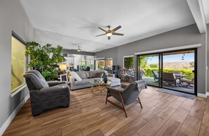 Ein großzügiger Wohnbereich mit einer Couch, Sesseln und einem Couchtisch. Ein großes Fenster bietet Blick auf Grünflächen und einen Balkon.