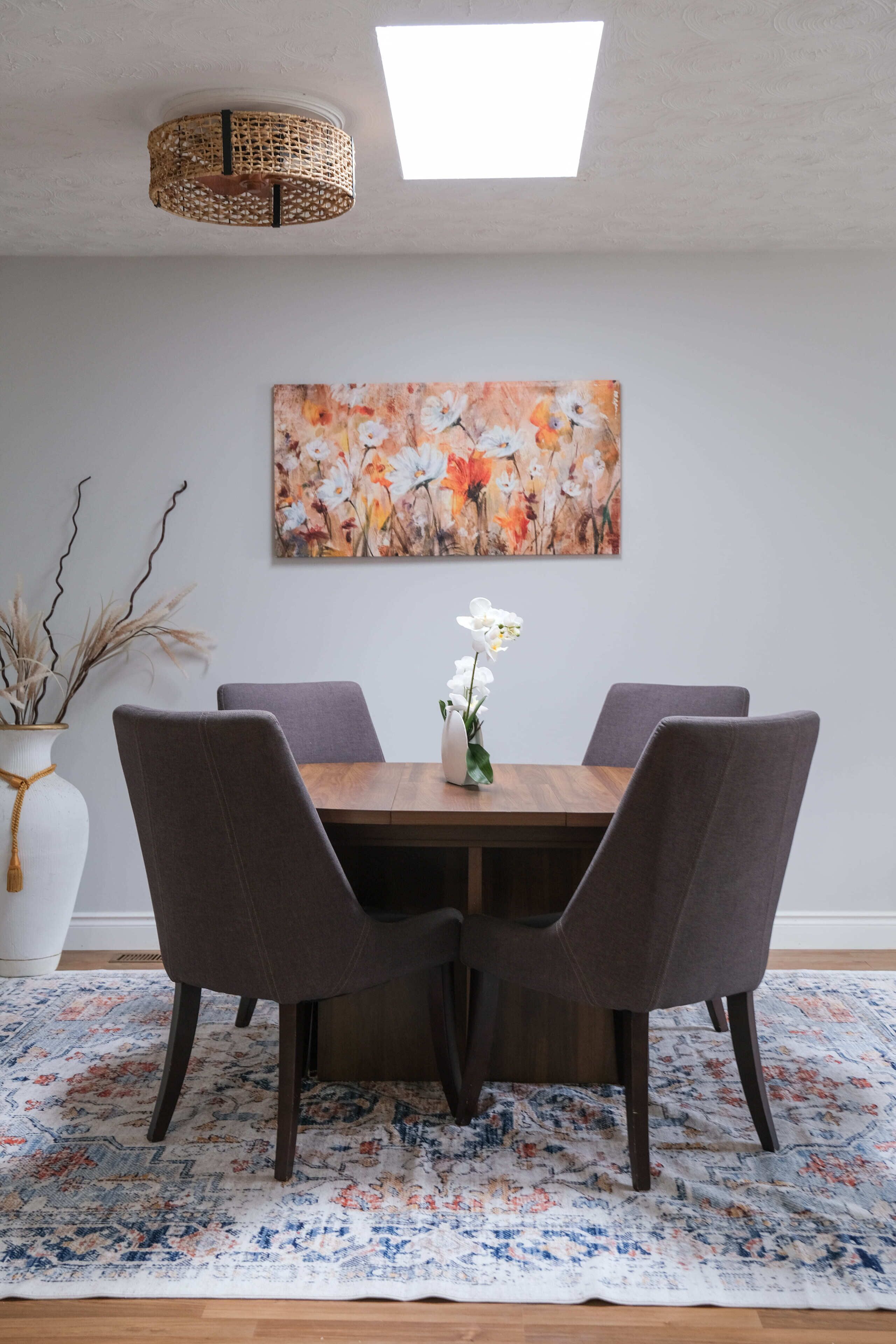 A dining area with a wooden table, four chairs, a vase with flowers, a painting on the wall, and a ceiling light fixture.
