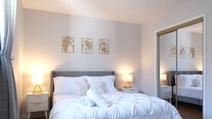 Ein ordentlich bezogenes Bett mit weißer Bettwäsche, zwei Nachttische mit Lampen, ein großer Spiegel und drei Wandbilder.