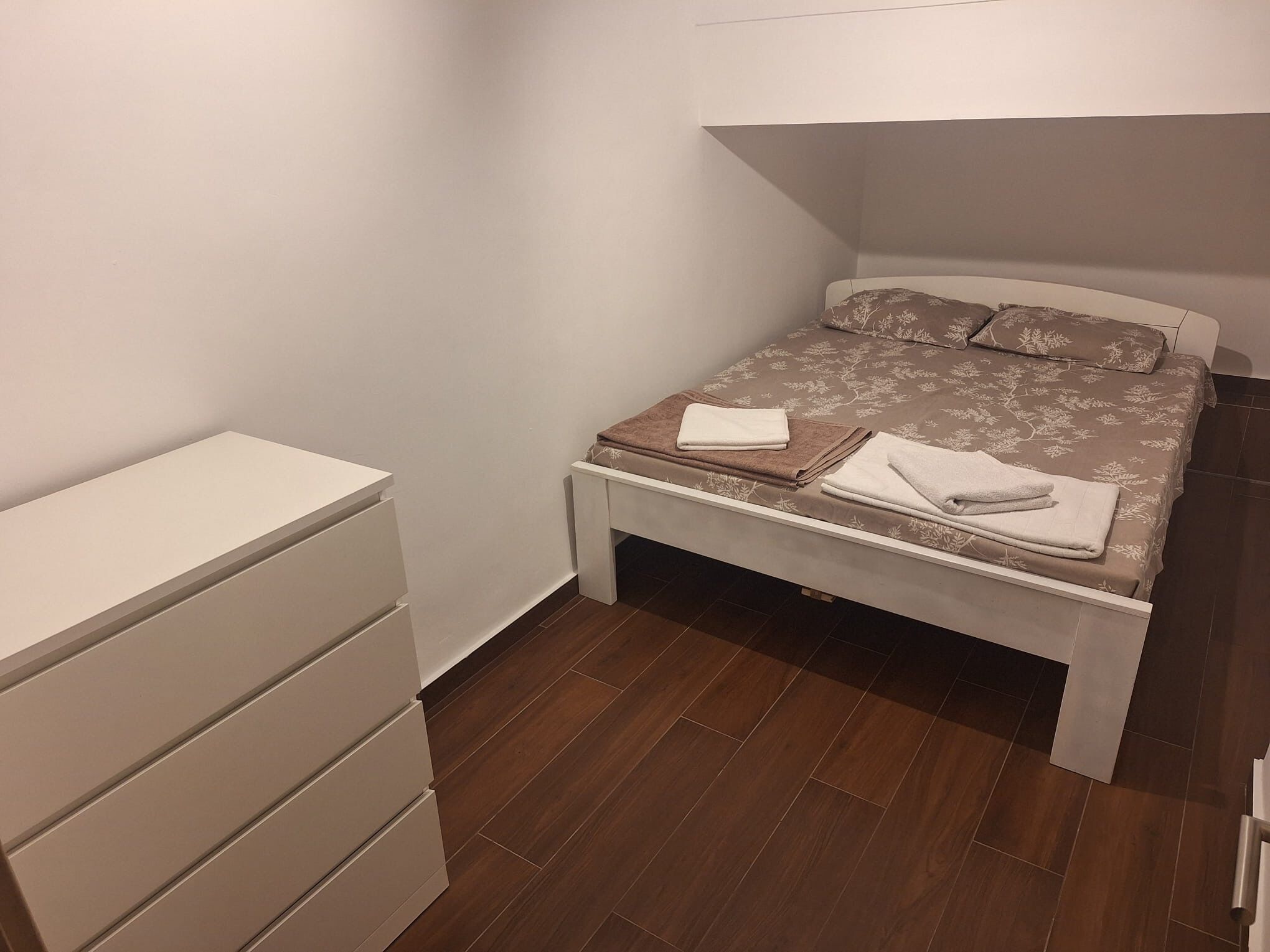 A bedroom with a bed, a bedside table, a dresser, and a small table with towels.