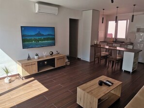Ett modernt vardagsrum med en platt-TV, trämöbler, en matplats och ett kök.