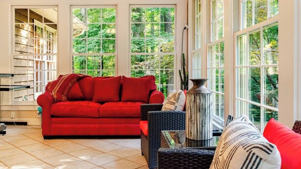 Una habitación iluminada por el sol con un sofá rojo, muebles de mimbre y ventanas amplias.