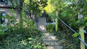 Eine steinerne Treppe, umgeben von dichtem Grün, führt zu einem Gebäude mit einem Metallgeländer.