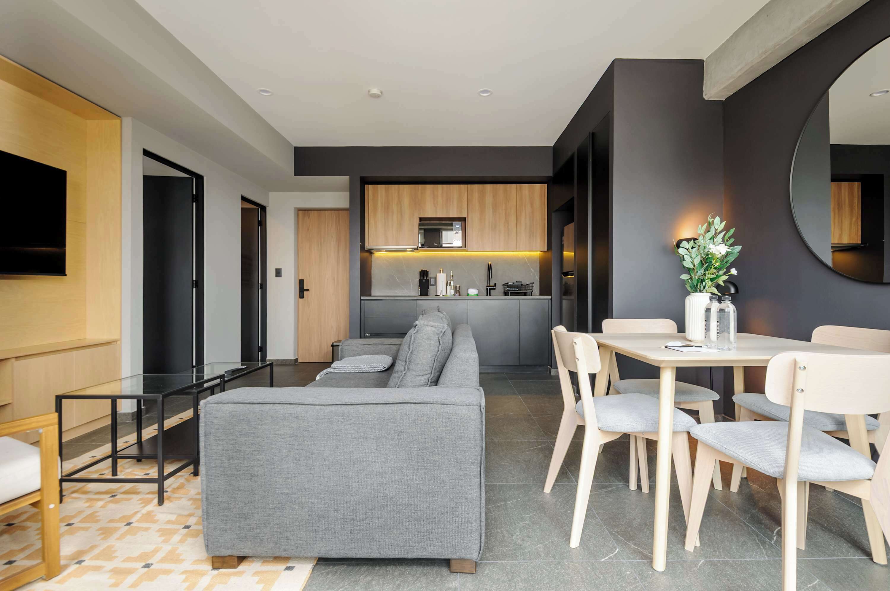 Ein modernes Wohnzimmer mit grauem Sofa, einem Esstisch mit Stühlen, einer Küchenzeile mit Holzschränken und einem an der Wand montierten Flachbildfernseher.
