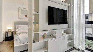 Een moderne hotelkamer met een bed, een televisie op een wit plankje, een nachtkastje met een lamp, en uitzicht door een raam.