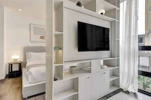 A modern hotel room with a bed, a TV on a white shelf, a bedside table with a lamp, and a view through a window. - w* | Luxury & Cozy Loft in Chapinero (Bogotá)