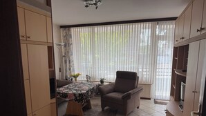 A room with a sofa, armchair, a small table with a floral tablecloth, a potted plant, and a window with sheer curtains.