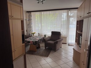 Quarto com sofá, poltrona, mesa pequena com toalha de mesa florida, planta em vaso e janela com cortinas translúcidas.