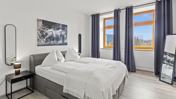 A modern bedroom with a bed, a nightstand, a mirror, a window with curtains, and a painting on the wall. - Faraway Homes Studios St.Michael (Liesingtal)