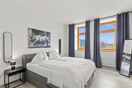 A modern bedroom with a bed, a nightstand, a mirror, a window with curtains, and a painting on the wall.