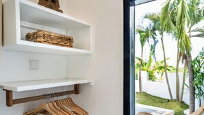 A room with a white wall, a wooden coat rack, a white shelf with a woven basket, and a view of a garden with palm trees. - Villa AMARÉ New Stylish 3BR Canggu Large Pool,
Cinema & BBQ (Badung)