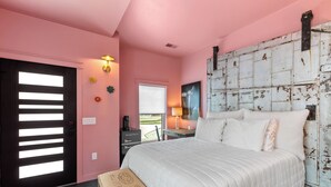 Ein Schlafzimmer mit einem Bett, einer Bank, einem Schreibtisch, einem Sessel, einer Tür mit Jalousien und einer Wandleuchte.