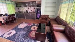 A living room with a brown leather sofa, a glass coffee table, a floral rug, and a kitchen in the background. - Full Furnished 2BHK (Kathmandu)