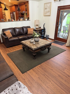 Ein Wohnzimmer mit braunem Ledersofa, einem Couchtisch mit Dekoration, einem Essbereich mit Holzsäcken und Blick durch eine Glastür.