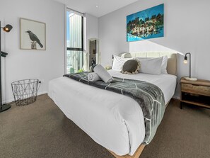 A modern bedroom with a large bed, a bedside table, a floor lamp, a wall-mounted painting, and a window with a view. - Onehunga Sanctuary I 2-Bed Apt. I Amazing Views (Grey Lynn)