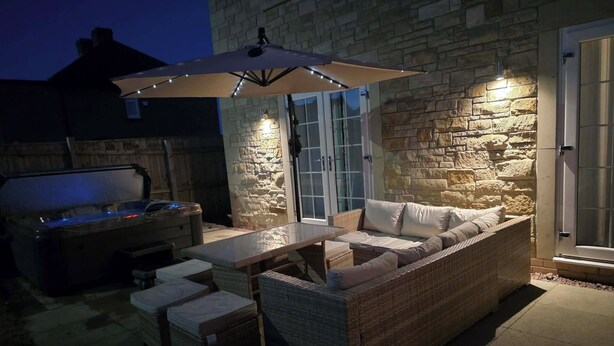An outdoor patio with a stone wall, a hot tub, a wicker sofa, and a table with an umbrella. - Rothbury House, Luxury Family Retreat with Hot tub and Playframe (Nortumberland)