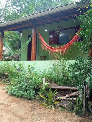 A rustic house with a green exterior, a hammock, and lush greenery around.