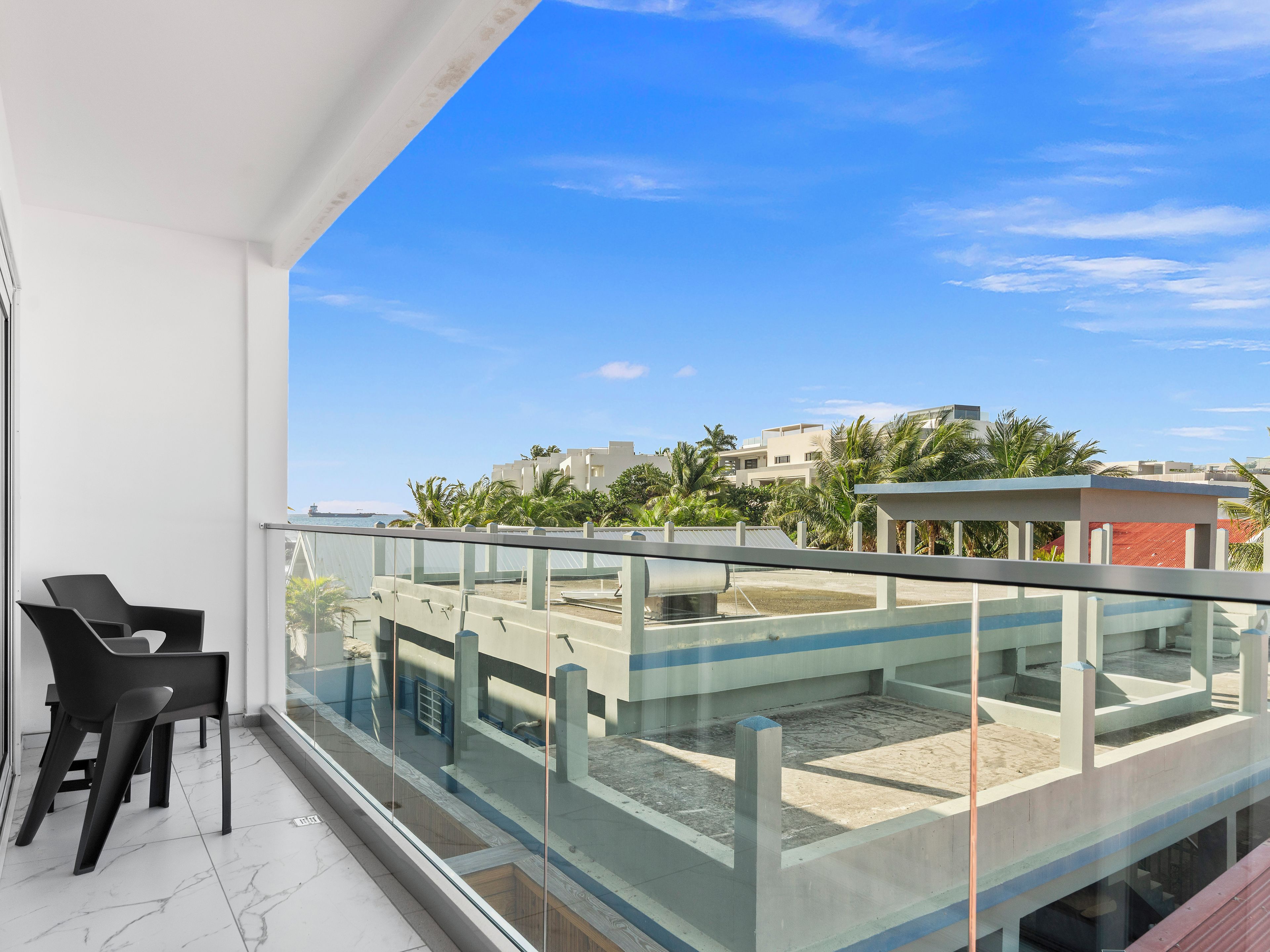 A balcony with a glass railing, a chair, and a view of buildings and palm trees.