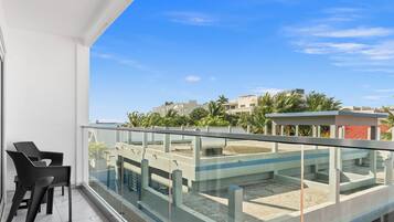 A balcony with a glass railing, a chair, and a view of buildings and palm trees.