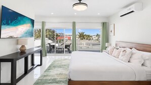 A modern hotel room with a large bed, a desk, a TV, and a balcony with a view of palm trees and buildings. - Nirvana - Stay Steps from the Sand in Simpson Bay (Simpson Bay)