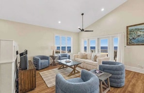 Ein geräumiges Wohnzimmer mit einer Couch, Sesseln, einem Couchtisch und einem Fernseher. Große Fenster bieten Blick auf das Meer.