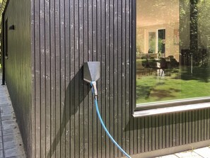 A building exterior with a vertical wooden panel design, a window with a view of a lawn and chairs, and a blue cable connected to a device on the wall. - 5 Star Holiday Home in Hals (Hals)