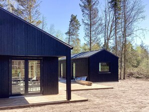 Eine moderne Hütte mit großen Fenstern, einer Holzterrasse und einem Metalldach, umgeben von Bäumen.