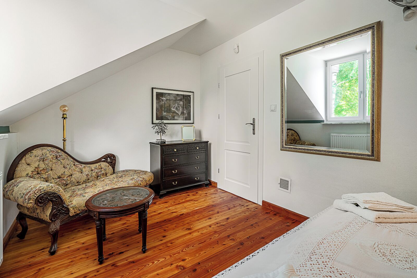 A room with a wooden floor, a floral sofa, a small round table, a dark dresser, a white door, and a large mirror.