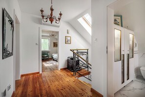 A room with a wooden floor, a chandelier, a staircase, and a view of a bedroom with a bed and a window.