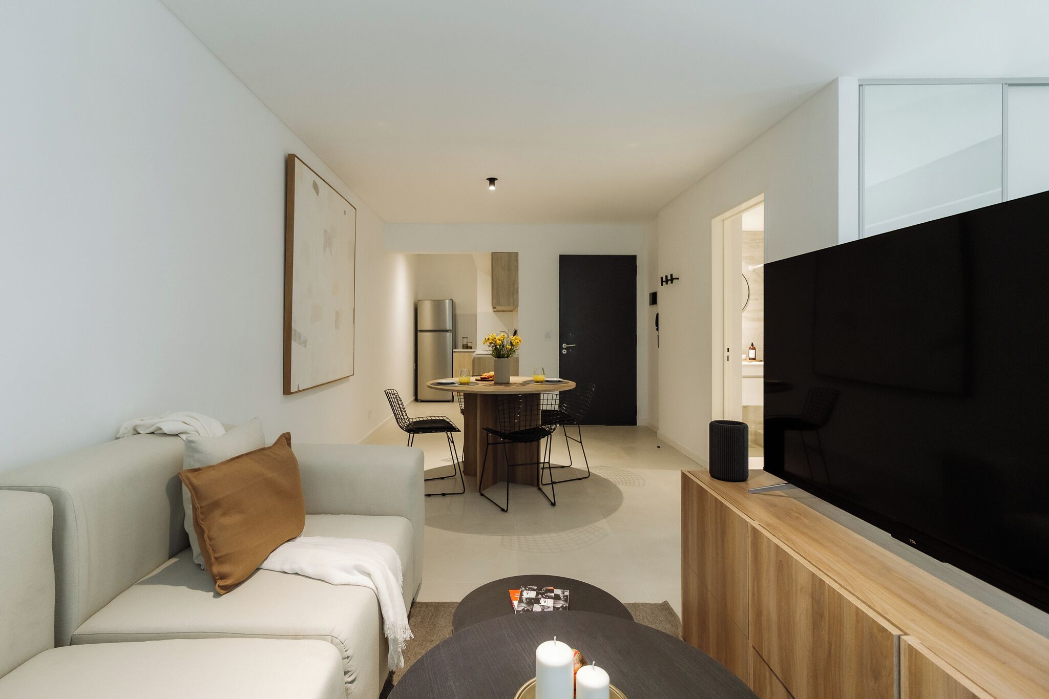 Una sala de estar moderna con un sofá, una mesa de centro, un televisor de pantalla plana, un comedor con una mesa y sillas, y una cocina al fondo.