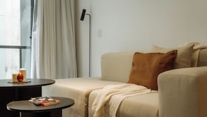 Una sala de estar moderna con un sofá, una mesita redonda con un libro, una lámpara de pie y una ventana con cortinas.