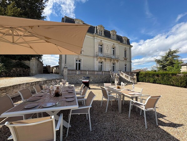 An outdoor dining area with tables and chairs, a large umbrella, and a building with classical architecture in the background. - Beautiful traditional French Chateau with heated pool & all modern comforts (Luxé)