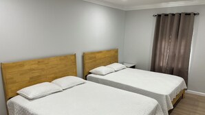 Una habitación con dos camas de madera, ropa de cama blanca, una mesita de noche pequeña y una ventana con cortinas.