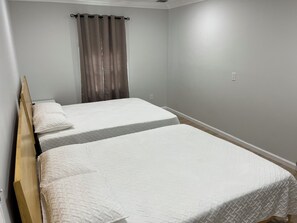 Une chambre avec deux lits simples, une tête de lit en bois, une fenêtre avec des rideaux et un interrupteur sur le mur.