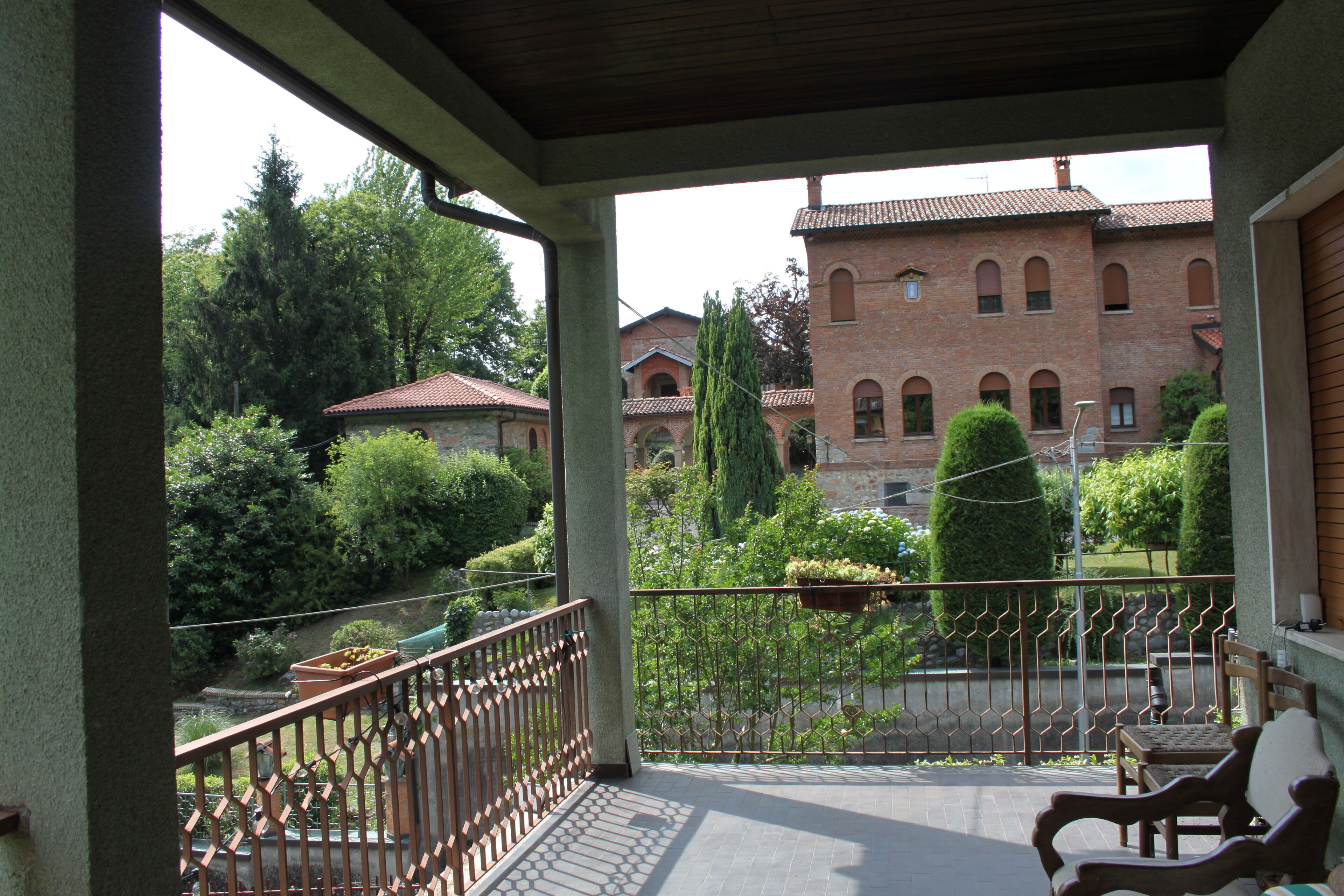 A covered balcony with a wrought iron railing, a wooden bench, and a view of a brick building with arched windows.