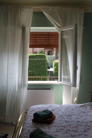 Un dormitorio con cama, ventana con cortinas y vista a un seto verde y un edificio de ladrillo.