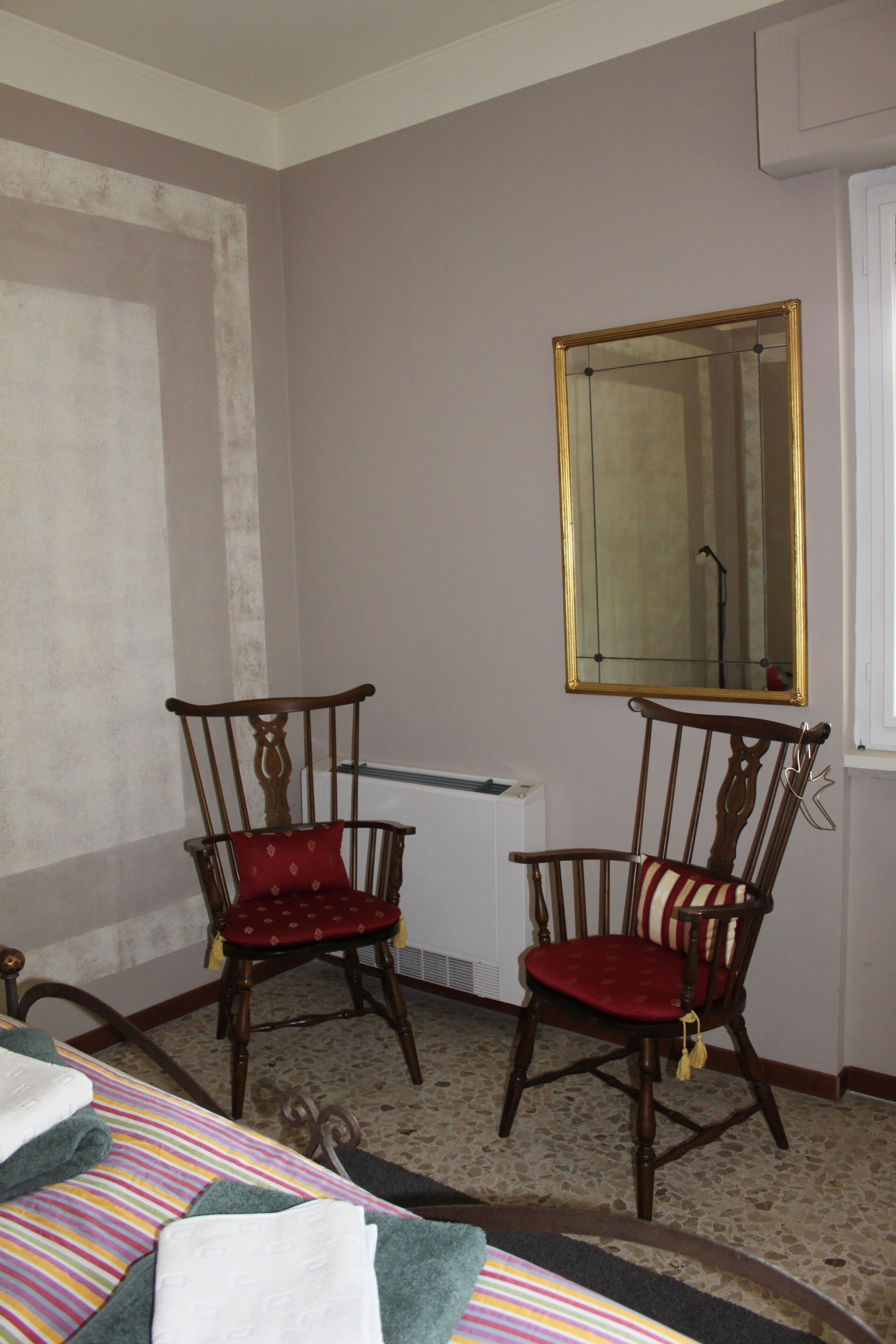 A room with two wooden chairs with red cushions, a bed with striped bedding, a large mirror on the wall, and a radiator.