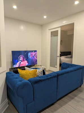 Ein Wohnzimmer mit einem blauen Sofa, einem Fernseher, der eine Sendung zeigt, und einem kleinen Tisch mit einem dekorativen Gegenstand.