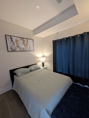 Ein Schlafzimmer mit einem Bett, einer Nachttischlampe, einem Wandbild und einem Fenster mit Vorhängen.