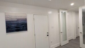 Un couloir avec un plancher en bois, des murs blancs et un tableau encadré représentant une scène de plage. On y trouve des portes blanches avec des panneaux de verre et un luminaire au plafond.