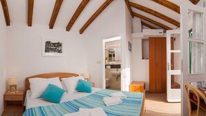 Ein Schlafzimmer mit Bett, Kissen, einem Nachttisch, einer Holzbalkendecke, einem durchgehenden Türspalt sichtbaren Badezimmer und einem Sessel.