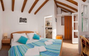 Ein Schlafzimmer mit Bett, Kissen, einem Nachttisch, einer Holzbalkendecke, einem durchgehenden Türspalt sichtbaren Badezimmer und einem Sessel.