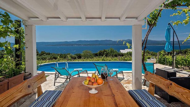 Ein Holztisch mit Obstschale, zwei Stühlen und Blick auf einen Swimmingpool und das Meer.