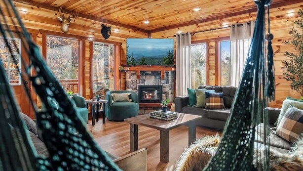 L’intérieur chaleureux d’un chalet avec une cheminée en pierre, des murs en bois et une vue sur la forêt grâce à de grandes fenêtres.