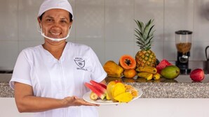 Una persona con uniforme blanco sostiene un plato de fruta en una cocina, con diversas frutas y una licuadora sobre la encimera.